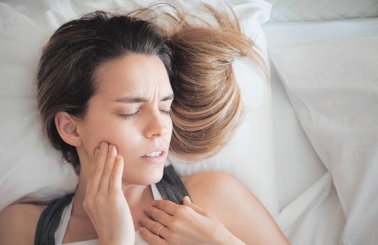 woman in bed with eyes closed and hand on jaw 